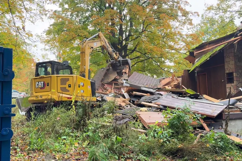 House demolition in progress with a roll-off dumpster beside the excavator
