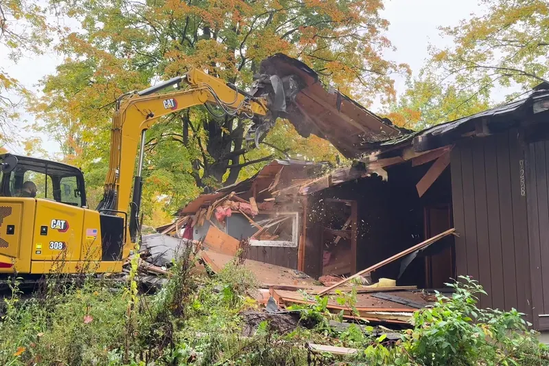 Excavator grapple lifting a roof section off a house being demolished
