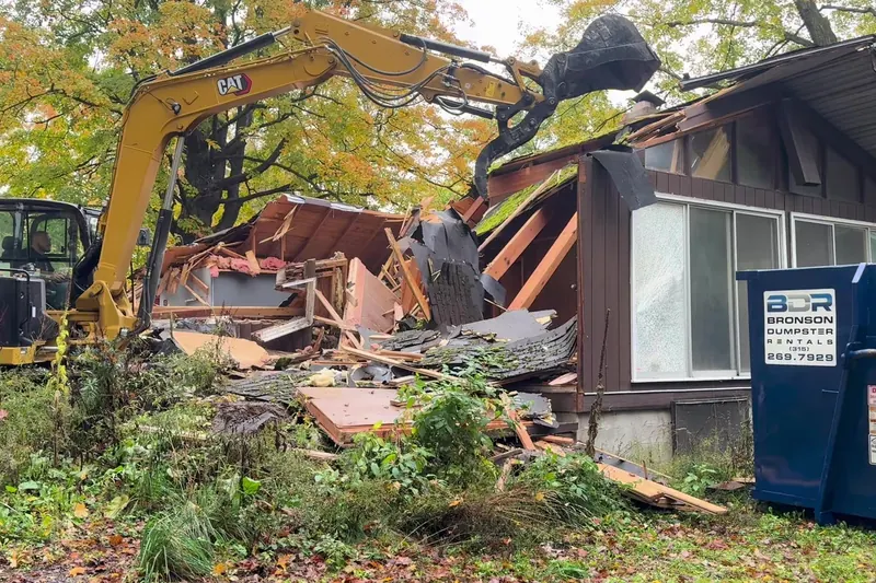 Excavator pulling the front wall of a house, roll-off dumpster on site